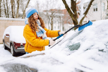 A woman in bright jacket clears snow from car windshield in morning in snowy area. Female driver...