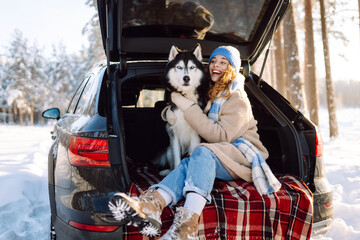 A happy woman and her pet sit on a blanket in the trunk of a car. A young woman in warm clothes with her husky enjoys a snowy day in the forest on a road trip. Concept of friendship, fun, adventure. © maxbelchenko