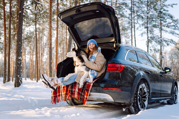 A happy woman and her pet sit on a blanket in the trunk of a car. A young woman in warm clothes...
