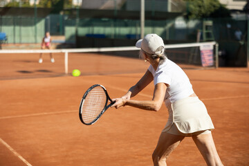 Rear view of focused european aged woman playing tennis match in open-air court of tennis club