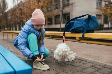 A beautiful, happy teenage girl, a gymnast or cheerleader, with pom-poms and a backpack, ties her shoelaces at a stadium outdoors. Photo, sports concept, cheerleading.