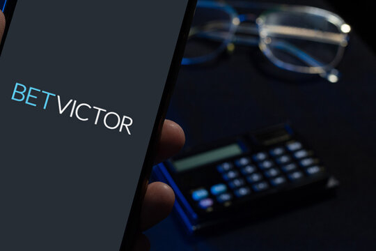 Representation of the BetVictor app on a smartphone screen held in one hand on a table with glasses and a calculator, concept of online sports betting