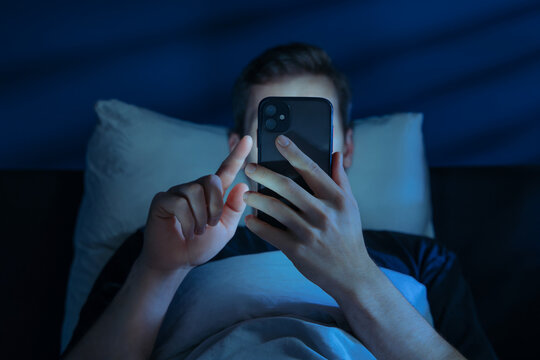 Close up of young man, alone in the dark bedroom, holds his smartphone close to face while scrolling social media or texting late at night. Screen dependence, excessive consumption of content