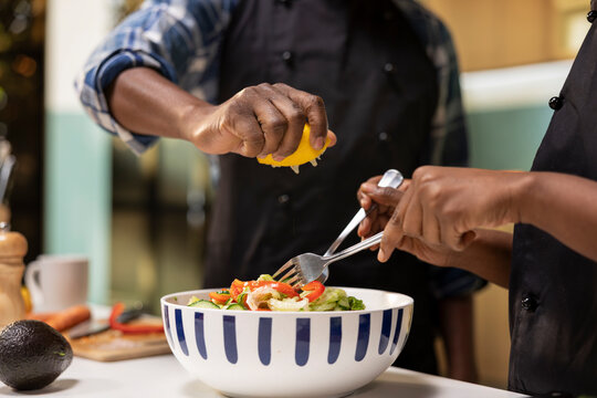 Young black couple adding lemon juice over salad and mixing goods in bowl, enjoying meal prep at home. Man and woman seasoning meal with organic fresh ingredients, daily lifestyle. - Powered by Adobe
