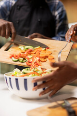 Close up of black person adding freshly cut veggies in a bowl to make delicious salad, having...