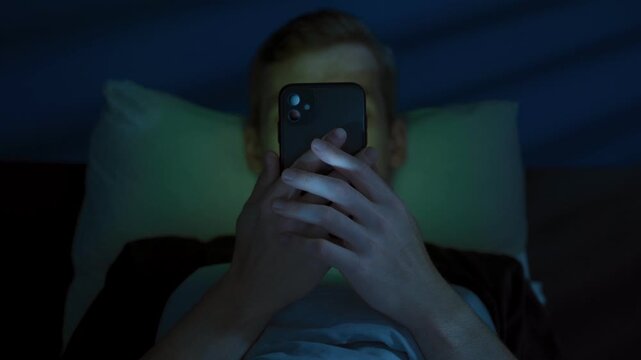 Close up of awake man lies in bed at night, holding his phone close to his face, illuminated by the bright colored light of the screen, deeply immersed in consuming content, addicted to social media. 