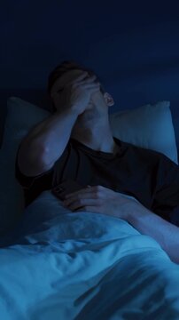 Tired young man lies awake in a dark bedroom, doom scrolling social media or news feed on his smartphone late at night and covering his face in frustration and exhaustion. Insomnia. Vertical video