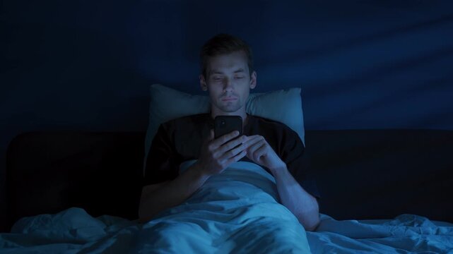 A tired man lies awake in a dark bedroom, staring at his glowing smartphone and doom scrolling late at night despite his growing fatigue. Digital addiction or insomnia concept. Copy space 