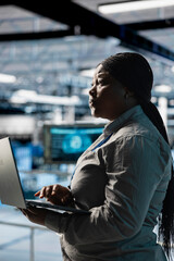 Woman in server farm using laptop, automating system diagnostics using machine learning predictions. Female technician in data center using notebook to configure gear for AI tasks