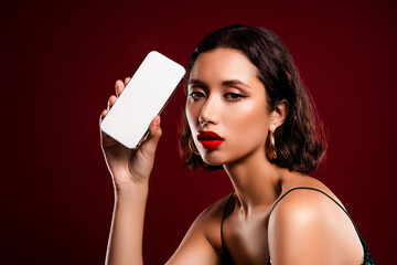 Luxurious young woman showcasing a smartphone against a rich red background, radiating beauty and...