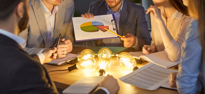 Cropped view of business team brainstorming around table with glowing light bulbs symbolizing creativity. Group of professionals analyzing pie chart, discussing business strategy together. Banner.
