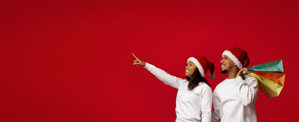 A happy couple wearing Santa hats stands against a bright red backdrop. They are holding colorful...