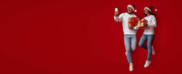 A joyful couple dressed in festive attire stands against a red backdrop, holding gifts and taking a...