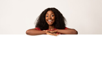 A happy young African American woman is leaning against a large blank advertisement board. She is...