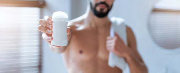 A man stands in a bright bathroom holding a stick of deodorant. He is shirtless and has a towel...