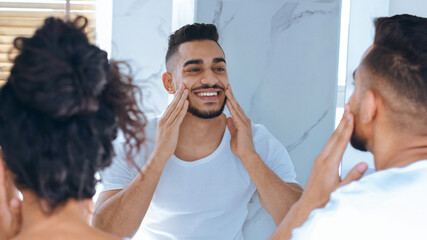 A man is enjoying his skincare routine in a bright bathroom. He smiles as he gently applies a...
