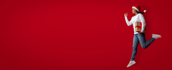 A person wearing a Santa hat jumps happily while holding a gift on a vibrant red background. The...