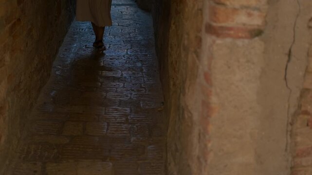 Woman walking in a narrow cobblestone alley. Young woman wearing a long dress and sandals walking through a dark, narrow cobblestone street in a historic european old town, emerging from the shadows