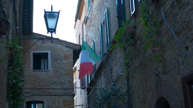 Italian flag waving in an alley of a medieval town in tuscany. Picturesque narrow street in a historic italian village, with an old stone building, a vintage street lamp, and the flag of italy