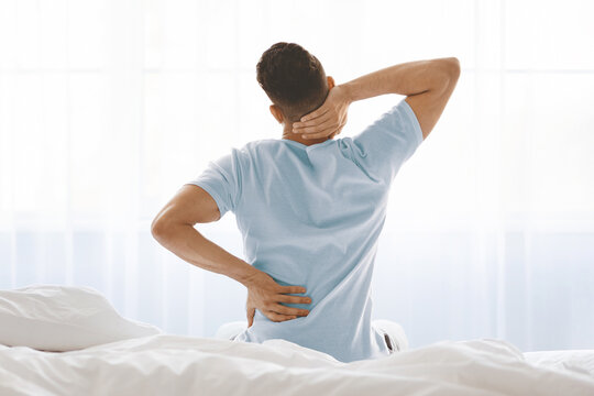 A man sits on a bed, holding his neck with one hand and his lower back with the other, showing signs of discomfort. Gentle morning light filters through the window, creating a calming atmosphere. - Powered by Adobe
