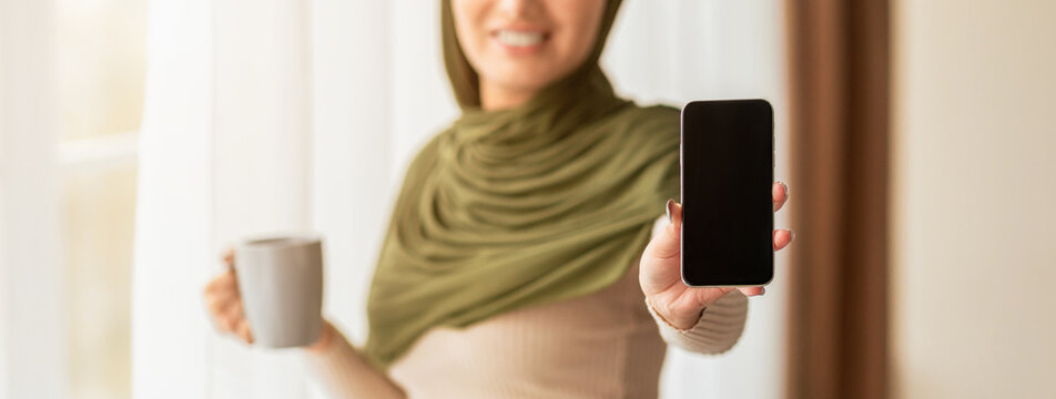 A woman wearing a green headscarf smiles while holding a cup of coffee in one hand and a smartphone in the other. Sunlight fills the cozy room, creating a warm atmosphere.