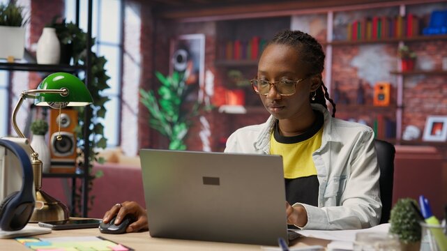 Teleworker analyzing data on laptop at desk and looking over paperwork, compiling analytical reports. Remote employee checking documents, using notebook to upload company information online, camera B