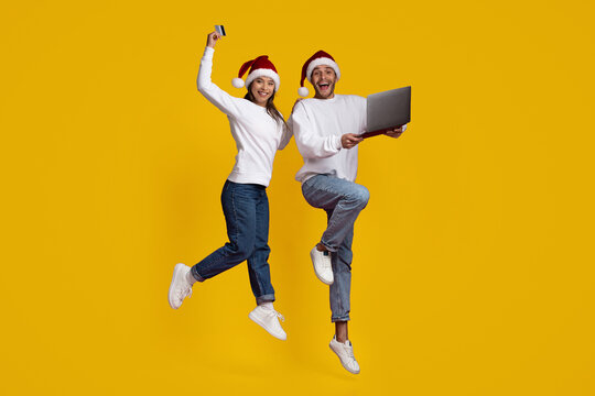 Two cheerful young adults wearing Santa hats jump against a bright yellow background while holding a credit card and a laptop, enjoying a fun holiday shopping experience. - Powered by Adobe
