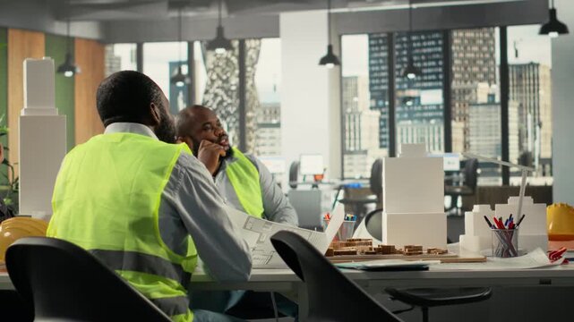 Property developers and architects in meeting reviewing blueprints and structural designs for zoning regulations. Team discusses project approval and city planning for housing growth. Camera B.