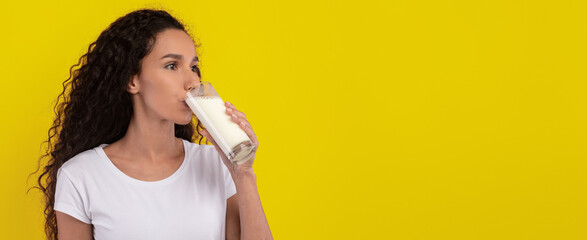 A woman with curly hair drinks a glass of milk, looking thoughtful. The vibrant yellow wall behind her adds a cheerful vibe to the scene, capturing a moment of simple pleasure.