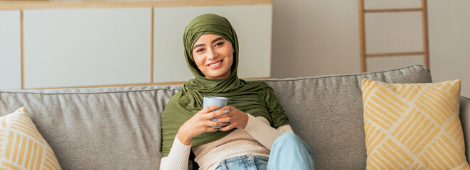 A young woman wearing a green scarf sits on a gray couch, holding a cup in her hands. She smiles...