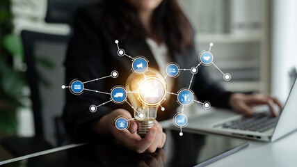 A businesswoman presents a glowing light bulb surrounded by digital icons, symbolizing innovation and connection in a modern workspace. Ideal for business themes. Tessel