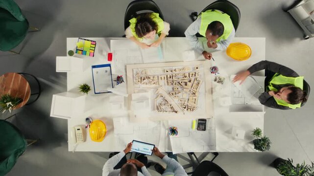 Top down view zoom in of real estate developers and architects in office reviewing blueprints and 3D models. Development team discusses urban planning and property investment strategies. Camera A.