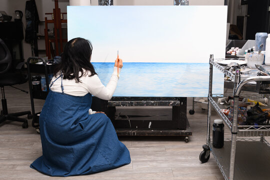 Female artist kneeling while painting a blue seascape on canvas - Powered by Adobe