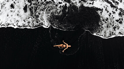 Aerial view of a woman floating peacefully on black sand beach, surrounded by foamy waves, capturing the essence of tranquility and relaxation in nature's embrace