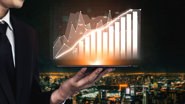 A businessman holds a laptop displaying a financial growth graph above a bustling city skyline at night, symbolizing success and investment strategies. Impute