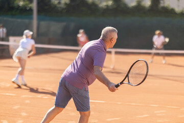 Active aged man hits tennis ball with racket in front of players in open-air court. Sportive elderly couple enjoying padel tennis