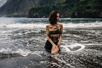 Fototapeta premium Young woman with wet hair kneeling in ocean waves on a rocky beach, wearing a green bikini, surrounded by lush cliffs and dramatic coastal scenery, embodying freedom and nature