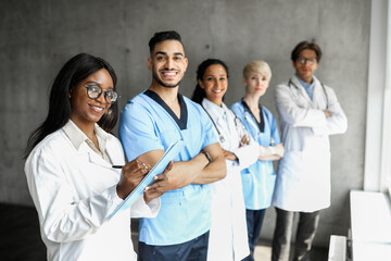 Fototapeta premium Cheerful international team of doctors having morning breefing, male and female medical workers standing by window, chatting, african american woman therapist holding medical chart, taking notes