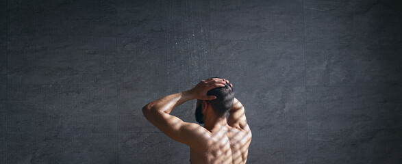 A man stands under a shower, water cascading down, in a softly lit space. The textured walls create an intimate atmosphere, highlighting the moment of relaxation and cleansing.