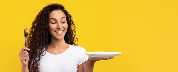 A young woman with curly hair holds a clean white plate in one hand and a fork in the other. She smiles joyfully, ready to enjoy her food in a vibrant setting.