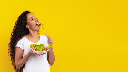 A young woman with long curly hair is holding a bowl of fresh salad and smiling as she takes a bite...