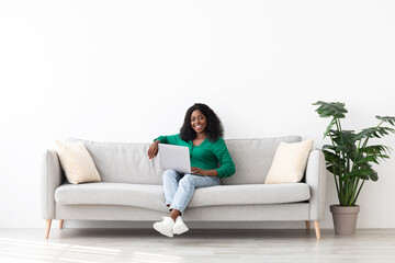 Obraz premium A woman sits comfortably on a gray sofa, smiling while using a laptop. The room features light colors and indoor plants, creating a cozy atmosphere.