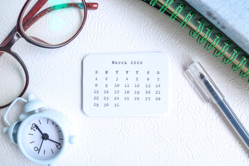 March 2026 paper sticker on a desk along with books, glasses, a clock, and a pen. March 2026 sticker on the desk