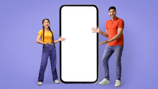 Two young adults are standing together, smiling broadly as they hold a large blank smartphone.
