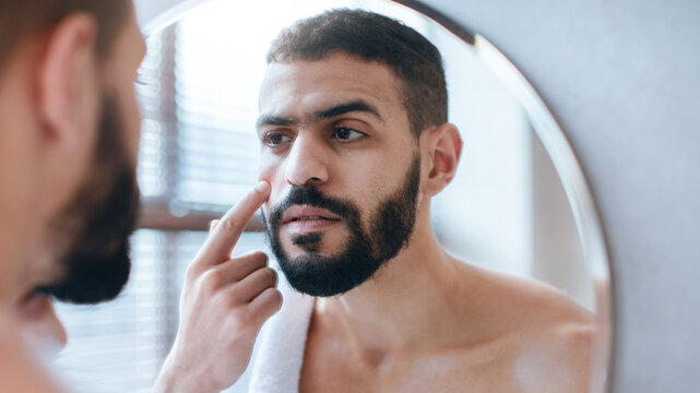 A man stands in front of a mirror, examining his face as he applies a skincare product. Sunlight filters through the window, creating a calm morning atmosphere.