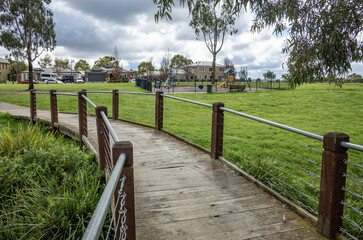 Fototapeta premium A wooden pedestrian bridge or boardwalk in a local park in Tarneit, Melbourne Australia, featuring cable-rail fencing, green open space, and a nearby playground with residential houses in background