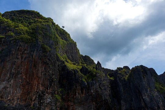 View of dramatic cliffs rising sharply, adorned with vibrant green foliage against a backdrop of cloudy skies, Phi Phi Island, Krabi, Thailand.