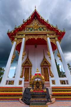 View of a Thai temple exhibiting ornate gold detailing contrasting against the white pillars and a stormy sky above, Phuket, Phuket, Thailand.