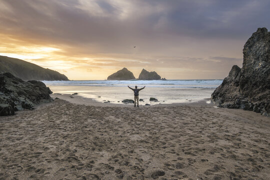 View of a lone figure stands silhouetted against the shimmering horizon, arms outstretched to embrace the golden light, nestled between rocky outcrops, Newquay, England, United Kingdom.