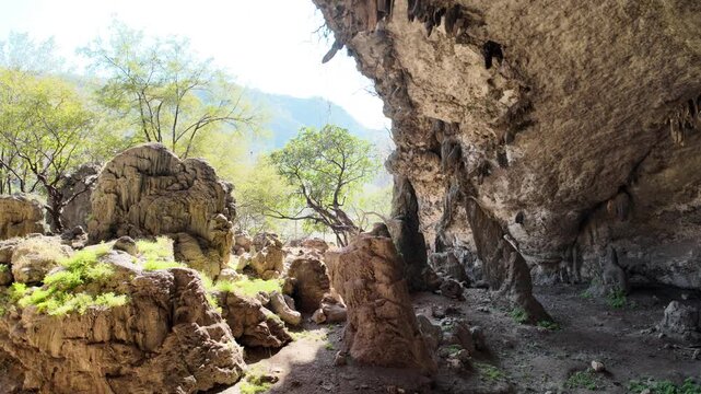 Natural cave entrance forming an arch, looking out towards a sunny, green landscape with trees and rugged hills under a clear blue sky in Dhofar, Oman, offering a sense of exploration